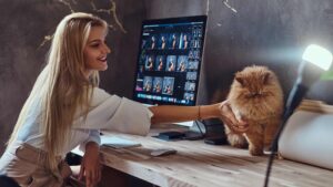 Blonde, female working at the computer and happily petting her pet cat.