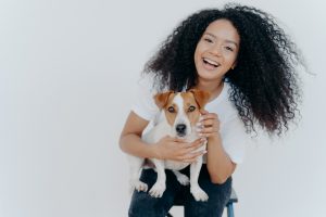 Portrait of joyful curly girl petting her dog, rejoicing buying jack russell terrier, smiles broadly, plays with animal, wears casual clothing, isolated over white background, enjoys good day.